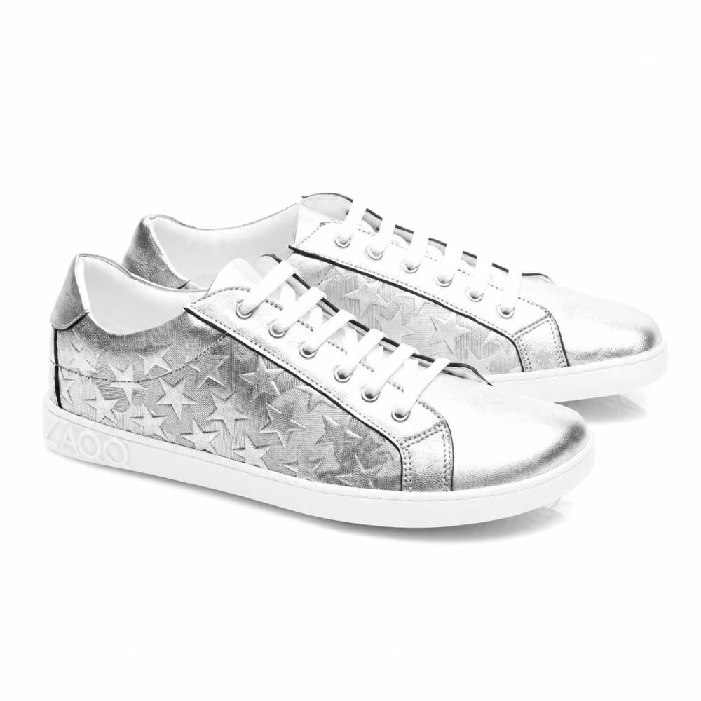 The SLOQ Stars Silver Sneakers from ZAQQ feature white laces and star patterns on the sides, complemented by a white sole with VAOO embossing at the heel. These shoes are characterized by a sleek and modern design with a shiny metallic finish.