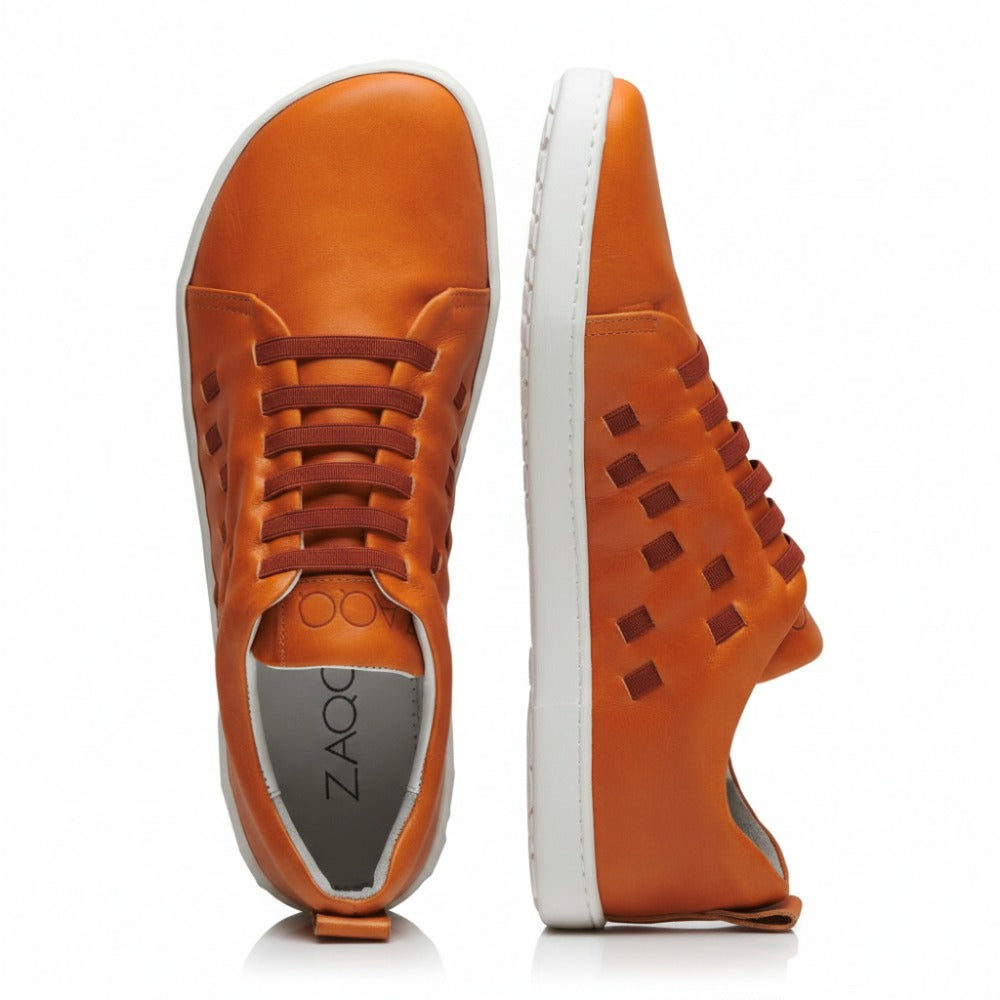 The STRAQ Sierra sneakers from ZAQQ are made of orange nappa leather and have white soles. They are characterized by striking side cutouts and a lace-up design. One sneaker is presented upright while the other lies flat, making the ZAQQ-Branding is highlighted on both the insole and the outer edge of the sole.